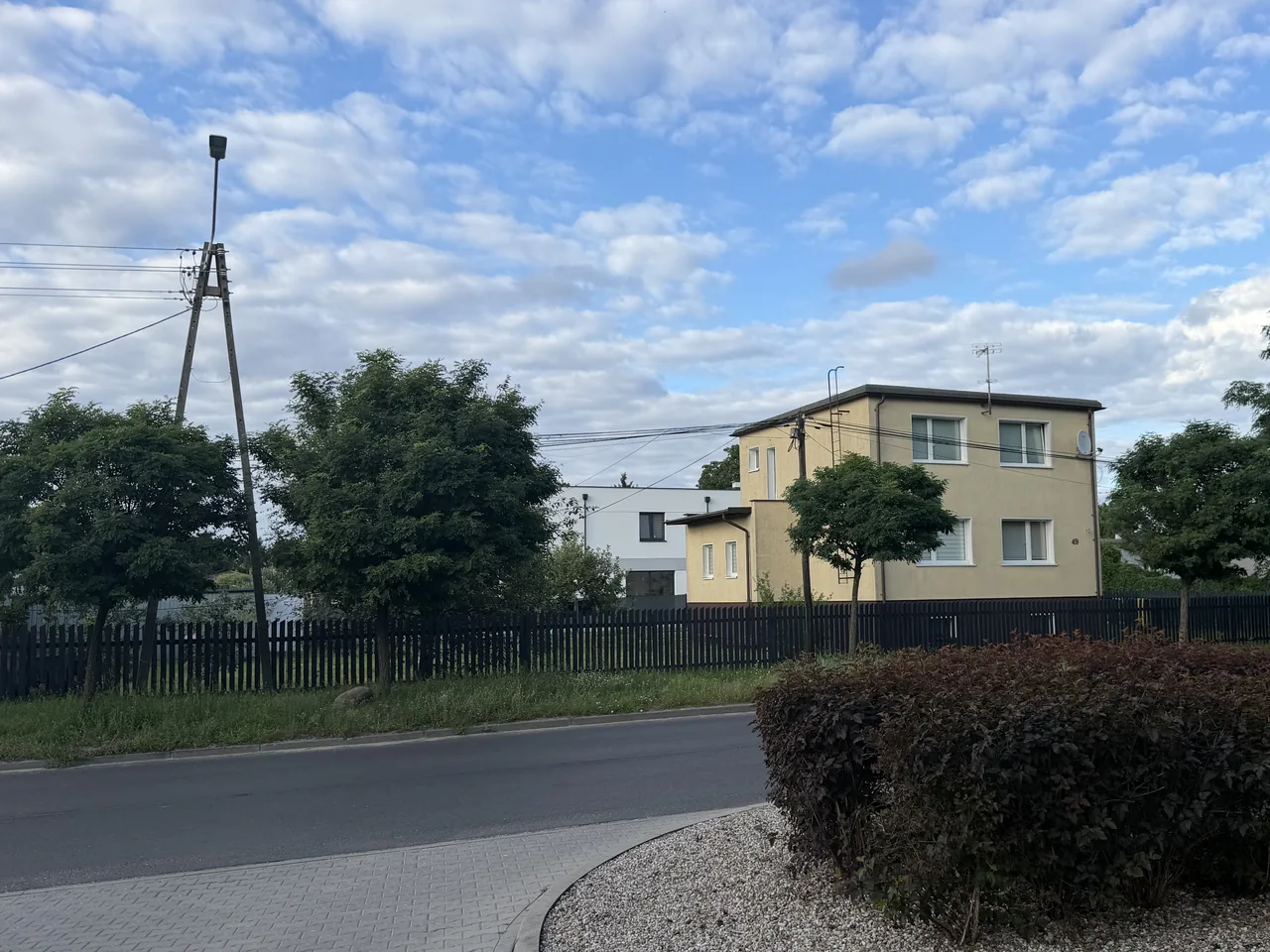 Skrzyżowanie w Białych Błotach — piętrowy dom z lat 90 w tynku, za nim bryła domu współczesnego z płaskim dachem